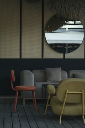 The image depicts a stylish indoor seating area with modern furniture. A round mirror hangs on a wall with a textured surface, reflecting an outdoor scene. The furniture includes an orange chair, a beige sofa with cushions, and a mustard-colored armchair, all set on wooden flooring. The decor is minimalistic with a muted and elegant color palette.