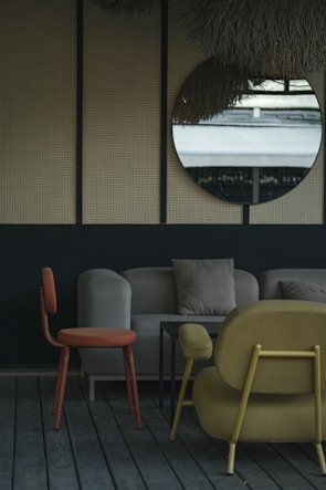 The image depicts a stylish indoor seating area with modern furniture. A round mirror hangs on a wall with a textured surface, reflecting an outdoor scene. The furniture includes an orange chair, a beige sofa with cushions, and a mustard-colored armchair, all set on wooden flooring. The decor is minimalistic with a muted and elegant color palette.