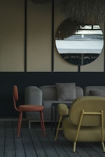 The image depicts a stylish indoor seating area with modern furniture. A round mirror hangs on a wall with a textured surface, reflecting an outdoor scene. The furniture includes an orange chair, a beige sofa with cushions, and a mustard-colored armchair, all set on wooden flooring. The decor is minimalistic with a muted and elegant color palette.