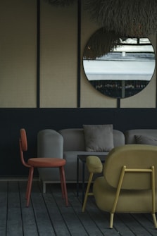 The image depicts a stylish indoor seating area with modern furniture. A round mirror hangs on a wall with a textured surface, reflecting an outdoor scene. The furniture includes an orange chair, a beige sofa with cushions, and a mustard-colored armchair, all set on wooden flooring. The decor is minimalistic with a muted and elegant color palette.