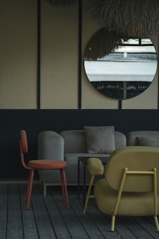 The image depicts a stylish indoor seating area with modern furniture. A round mirror hangs on a wall with a textured surface, reflecting an outdoor scene. The furniture includes an orange chair, a beige sofa with cushions, and a mustard-colored armchair, all set on wooden flooring. The decor is minimalistic with a muted and elegant color palette.