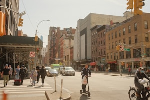 Pedestrians and cyclists move through a busy city intersection on a sunny day, with people crossing the street and a person riding a scooter. Surrounding buildings, including retail stores and apartments, reflect a typical urban environment.