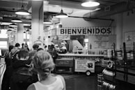 A black and white market setting with several people lined up at a food stall decorated with the word 'Bienvenidos'. The stall offers Mexican cuisine with items like tacos and breakfast burritos visible on signs. Industrial light fixtures hang from the ceiling, and there are various signs and menus displayed.