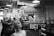 A black and white market setting with several people lined up at a food stall decorated with the word 'Bienvenidos'. The stall offers Mexican cuisine with items like tacos and breakfast burritos visible on signs. Industrial light fixtures hang from the ceiling, and there are various signs and menus displayed.