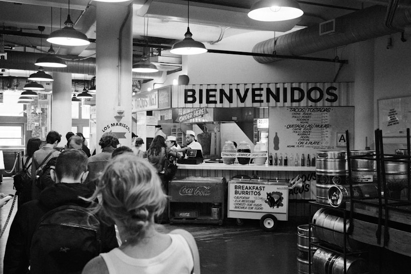 A black and white market setting with several people lined up at a food stall decorated with the word 'Bienvenidos'. The stall offers Mexican cuisine with items like tacos and breakfast burritos visible on signs. Industrial light fixtures hang from the ceiling, and there are various signs and menus displayed.