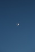 A crescent moon is positioned against a dark blue sky, accompanied by a bright star or planet nearby. The celestial objects stand out sharply against the vast, clear backdrop.