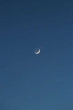A crescent moon is positioned against a dark blue sky, accompanied by a bright star or planet nearby. The celestial objects stand out sharply against the vast, clear backdrop.