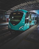 A modern, turquoise train is stationed inside a large, industrial-looking train depot with a high ceiling and metal beams. The train features sleek, aerodynamic design elements and is adorned with artistic graphics on its side.