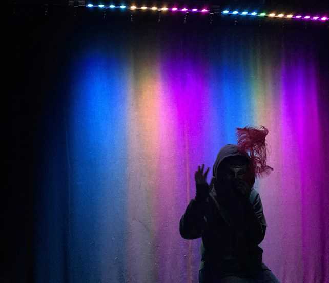 A dimly lit stage with a backdrop illuminated by a gradient of vibrant colors, including blue, green, pink, and purple. A person is partially visible in silhouette, wearing a hoodie and a mask or hat with feathers.