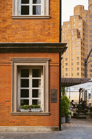 A red brick building with large white-framed windows is situated on a cobblestone street. A flower bed is positioned beneath the window, adding a touch of greenery. To the right, an outdoor cafe setup has people seated under two umbrellas. In the background, there is a taller, more modern beige building.