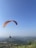A paraglider is soaring through a clear blue sky above a vast, green landscape with rolling hills and scattered small villages. The orange and red parachute contrasts with the serene sky and the expansive terrain below.