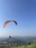 A paraglider is soaring through a clear blue sky above a vast, green landscape with rolling hills and scattered small villages. The orange and red parachute contrasts with the serene sky and the expansive terrain below.