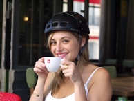A candid shot of a smiling woman wearing a black baseball cap embroidered with a subtle cross, holding a coffee cup.