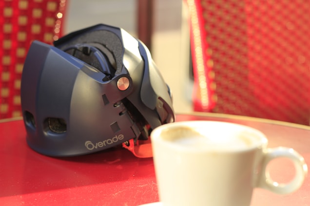 A folded bicycle helmet lies on a red table next to a steaming cup of coffee, with a background featuring red-patterned chairs. Sunlight casts soft shadows, emphasizing the helmet