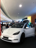 A white electric car is prominently displayed indoors, likely in a showroom or mall. The car's front door is open, and several people are gathered around, some appearing to be discussing or observing the vehicle. Overhead lighting and signage create a modern atmosphere.