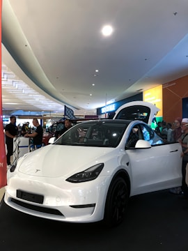 A white electric car is prominently displayed indoors, likely in a showroom or mall. The car's front door is open, and several people are gathered around, some appearing to be discussing or observing the vehicle. Overhead lighting and signage create a modern atmosphere.