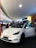 A white electric car is prominently displayed indoors, likely in a showroom or mall. The car's front door is open, and several people are gathered around, some appearing to be discussing or observing the vehicle. Overhead lighting and signage create a modern atmosphere.