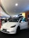 A white electric car is prominently displayed indoors, likely in a showroom or mall. The car's front door is open, and several people are gathered around, some appearing to be discussing or observing the vehicle. Overhead lighting and signage create a modern atmosphere.