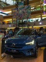 A Volvo C40 Recharge electric car is prominently displayed in a shopping mall. The vehicle is a glossy blue color and has its headlights turned on. Surrounding the car are informational displays, and a few people can be seen in the background. The mall interior is multi-leveled, with colorful decorations hanging from the ceiling, showcasing a vibrant and modern design.