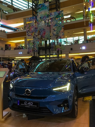 A Volvo C40 Recharge electric car is prominently displayed in a shopping mall. The vehicle is a glossy blue color and has its headlights turned on. Surrounding the car are informational displays, and a few people can be seen in the background. The mall interior is multi-leveled, with colorful decorations hanging from the ceiling, showcasing a vibrant and modern design.