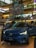 A Volvo C40 Recharge electric car is prominently displayed in a shopping mall. The vehicle is a glossy blue color and has its headlights turned on. Surrounding the car are informational displays, and a few people can be seen in the background. The mall interior is multi-leveled, with colorful decorations hanging from the ceiling, showcasing a vibrant and modern design.