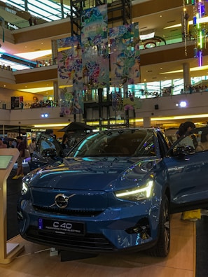 A Volvo C40 Recharge electric car is prominently displayed in a shopping mall. The vehicle is a glossy blue color and has its headlights turned on. Surrounding the car are informational displays, and a few people can be seen in the background. The mall interior is multi-leveled, with colorful decorations hanging from the ceiling, showcasing a vibrant and modern design.