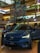 A Volvo C40 Recharge electric car is prominently displayed in a shopping mall. The vehicle is a glossy blue color and has its headlights turned on. Surrounding the car are informational displays, and a few people can be seen in the background. The mall interior is multi-leveled, with colorful decorations hanging from the ceiling, showcasing a vibrant and modern design.