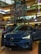 A Volvo C40 Recharge electric car is prominently displayed in a shopping mall. The vehicle is a glossy blue color and has its headlights turned on. Surrounding the car are informational displays, and a few people can be seen in the background. The mall interior is multi-leveled, with colorful decorations hanging from the ceiling, showcasing a vibrant and modern design.