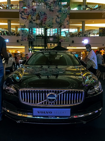 A sleek, black Volvo car is displayed prominently in a modern shopping mall setting. Several people are gathered around, likely admiring or inspecting the vehicle. The environment is bustling with activity, indicated by the presence of various storefronts and a multi-level structure in the background. Colorful banners and lighting reflections are visible on the car's glossy surface.
