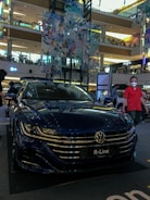 A shiny, dark blue Volkswagen R-Line car is displayed in a multi-level shopping mall. The car's front grille and headlights are clearly visible. The mall's upper levels are in the background, featuring various shops and illuminated signs. Decorative banners showcasing a floral and geometric design hang from above. There are people around the car; one individual in a red shirt and glasses stands on the right.