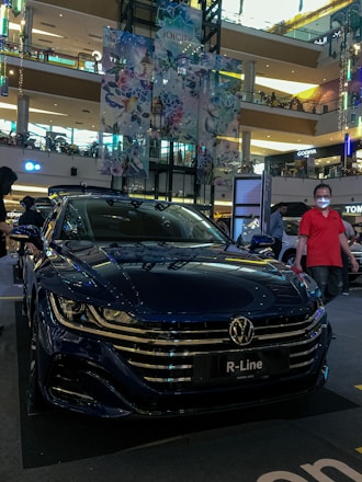 A shiny, dark blue Volkswagen R-Line car is displayed in a multi-level shopping mall. The car's front grille and headlights are clearly visible. The mall's upper levels are in the background, featuring various shops and illuminated signs. Decorative banners showcasing a floral and geometric design hang from above. There are people around the car; one individual in a red shirt and glasses stands on the right.