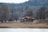 A small red house with a tiled roof is situated in a serene landscape. Surrounded by leafless trees and dry grass, the scene conveys a sense of quiet and solitude. The background features a forested hillside under a cloudy sky, indicating a winter or early spring setting.