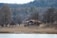 A small red house with a tiled roof is situated in a serene landscape. Surrounded by leafless trees and dry grass, the scene conveys a sense of quiet and solitude. The background features a forested hillside under a cloudy sky, indicating a winter or early spring setting.