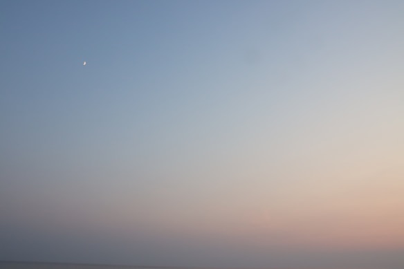 A serene sky transitions from a soft blue to pastel hues as the sun begins to set. A faint crescent moon is visible in the upper left corner, adding a subtle celestial element to the tranquil scene.