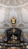 An ornate interior featuring a detailed stained glass window with a radiant eye motif. Below, a sculptural arrangement includes angelic figures and a central figure with a halo holding a sphere. Rich architectural details, gilded elements, and symbolic religious imagery are present.