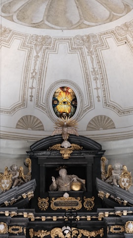 An ornate interior featuring a detailed stained glass window with a radiant eye motif. Below, a sculptural arrangement includes angelic figures and a central figure with a halo holding a sphere. Rich architectural details, gilded elements, and symbolic religious imagery are present.