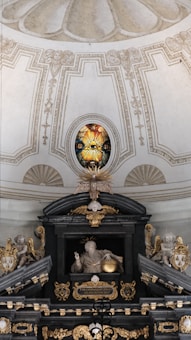 An ornate interior featuring a detailed stained glass window with a radiant eye motif. Below, a sculptural arrangement includes angelic figures and a central figure with a halo holding a sphere. Rich architectural details, gilded elements, and symbolic religious imagery are present.