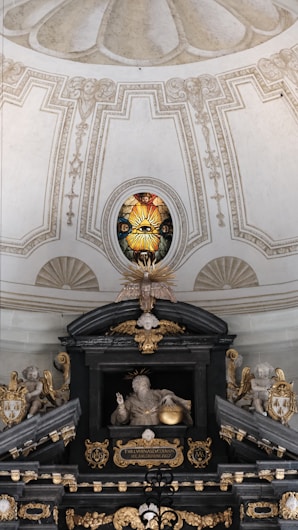 An ornate interior featuring a detailed stained glass window with a radiant eye motif. Below, a sculptural arrangement includes angelic figures and a central figure with a halo holding a sphere. Rich architectural details, gilded elements, and symbolic religious imagery are present.