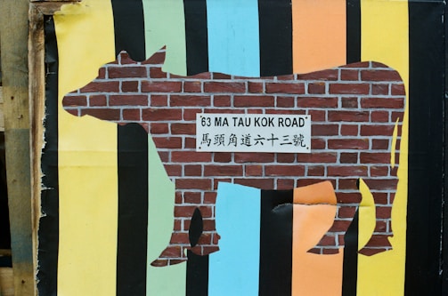 A silhouette of a cow is filled with a brick wall texture and features a sign in its center reading '63 MA TAU KOK ROAD' along with Chinese characters. The background consists of vertical stripes in various colors including yellow, blue, black, and orange.