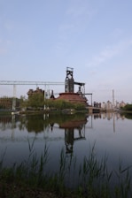 A large industrial plant with tall smokestacks is reflected in a calm body of water, surrounded by greenery and a blue sky.