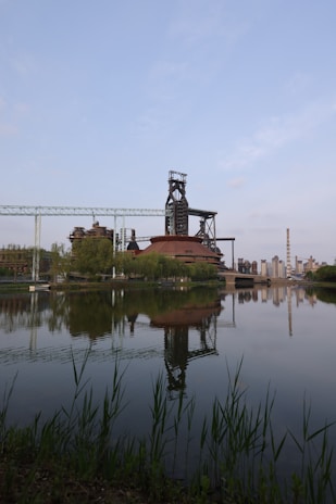 A large industrial plant with tall smokestacks is reflected in a calm body of water, surrounded by greenery and a blue sky.