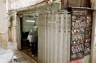An older man is assisting another person into a small, narrow shop or room with partially open metal sliding doors. The walls are adorned with graffiti, and there are various signs with writing inside the shop. The setting feels like an urban alleyway, and the overall environment appears worn yet lively.