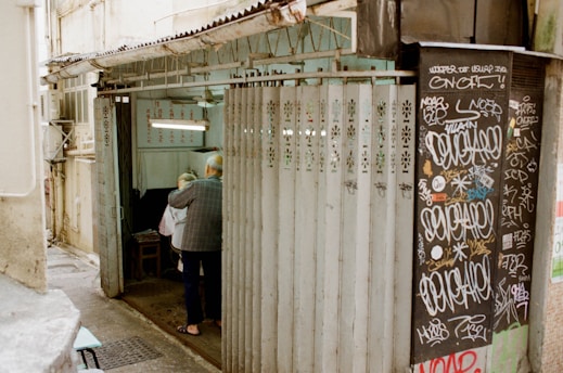 An older man is assisting another person into a small, narrow shop or room with partially open metal sliding doors. The walls are adorned with graffiti, and there are various signs with writing inside the shop. The setting feels like an urban alleyway, and the overall environment appears worn yet lively.