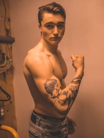 A shirtless young man with a muscular build poses while flexing his arm to display a prominent tattoo. The tattoo features a woman in a sailor's hat and nautical themes. The lighting is warm, creating a sepia-toned effect, and the setting appears to be indoors with visible wires and a wooden panel in the background.