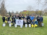 A group of people is gathered outdoors in a park, holding trash pickers, buckets, and bags for a cleanup event. They are wearing casual and some are in protective gloves, with the background showing trees and a pavilion. A large banner displaying 'Earth Day' is visible in the front.