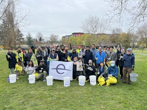 A group of people is gathered outdoors in a park, holding trash pickers, buckets, and bags for a cleanup event. They are wearing casual and some are in protective gloves, with the background showing trees and a pavilion. A large banner displaying 'Earth Day' is visible in the front.