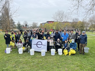 A group of people is gathered outdoors in a park, holding trash pickers, buckets, and bags for a cleanup event. They are wearing casual and some are in protective gloves, with the background showing trees and a pavilion. A large banner displaying 'Earth Day' is visible in the front.