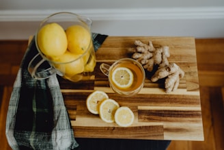 A wooden cutting board displays a glass mug filled with tea and a lemon slice. Next to the mug, fresh ginger root is placed alongside sliced lemons. A glass pitcher filled with whole lemons and a green plaid cloth are also present, contributing to a rustic kitchen setting.