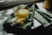 A glass cup filled with tea and a slice of lemon is placed on a dark green patterned cloth. The scene is set by a window with natural light casting shadows, creating a calm and tranquil atmosphere.
