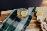 A clear glass cup filled with tea and a lemon slice on a wooden surface. Beside the cup, there is a dark green plaid cloth. To the right, there are three slices of lemon arranged next to a piece of fresh ginger.